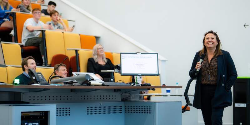 Lecturer stood in front of computer and tiered lecture theatre seating.